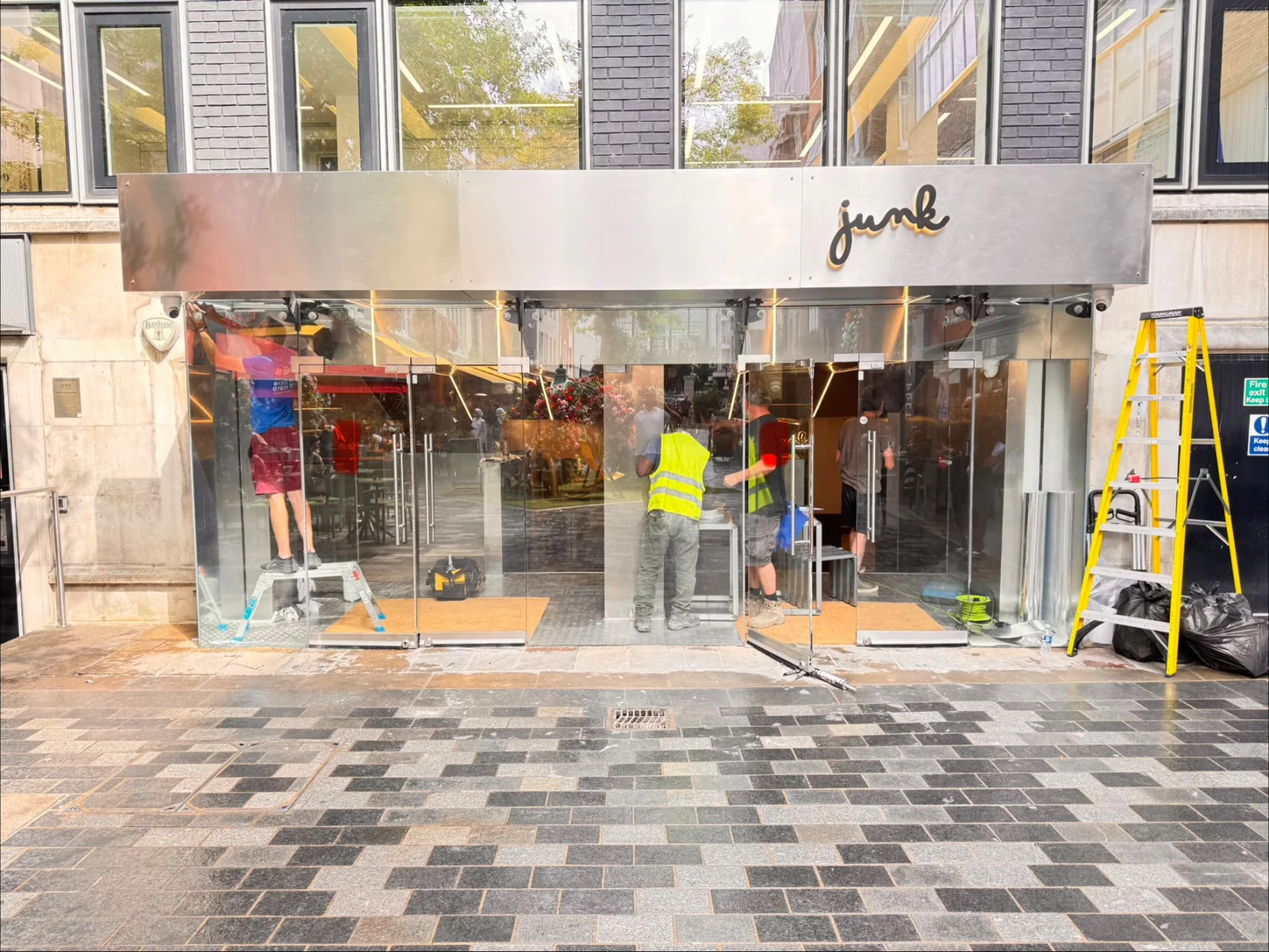 Brushed metal Junk fascia with script lettering above a glazed shopfront, with fitters working at the entrance