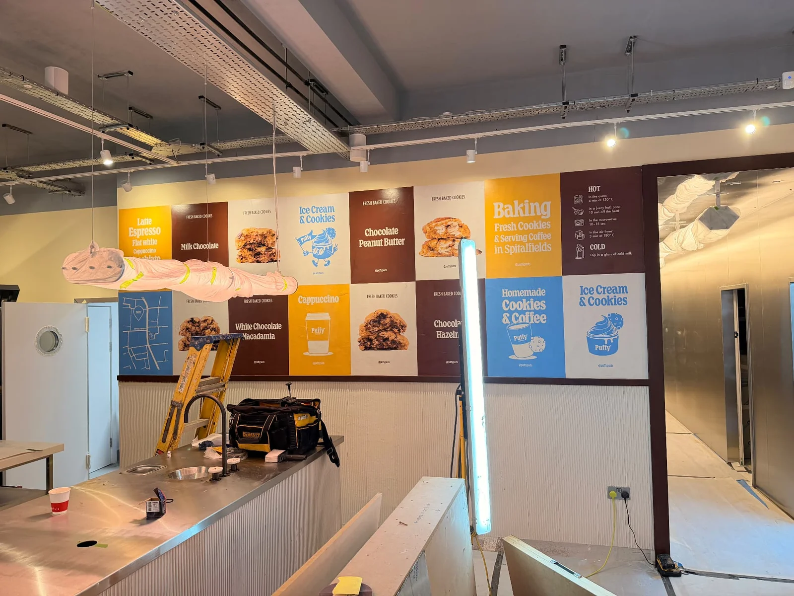 Grid of Puffy printed wall posters above a stainless steel counter, including a panel reading “in Spitalfields”