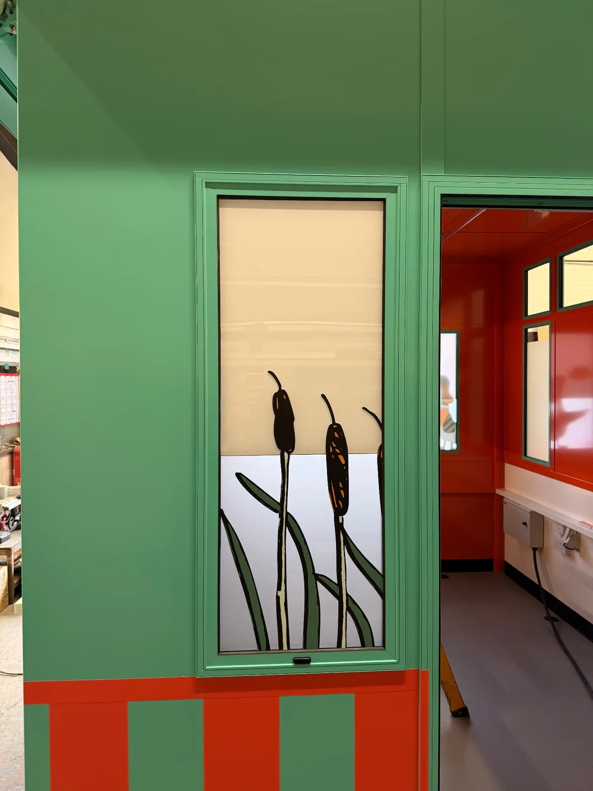 Close-up of an illustrated window covering showing bulrushes on a frosted panel in the kiosk’s green frame
