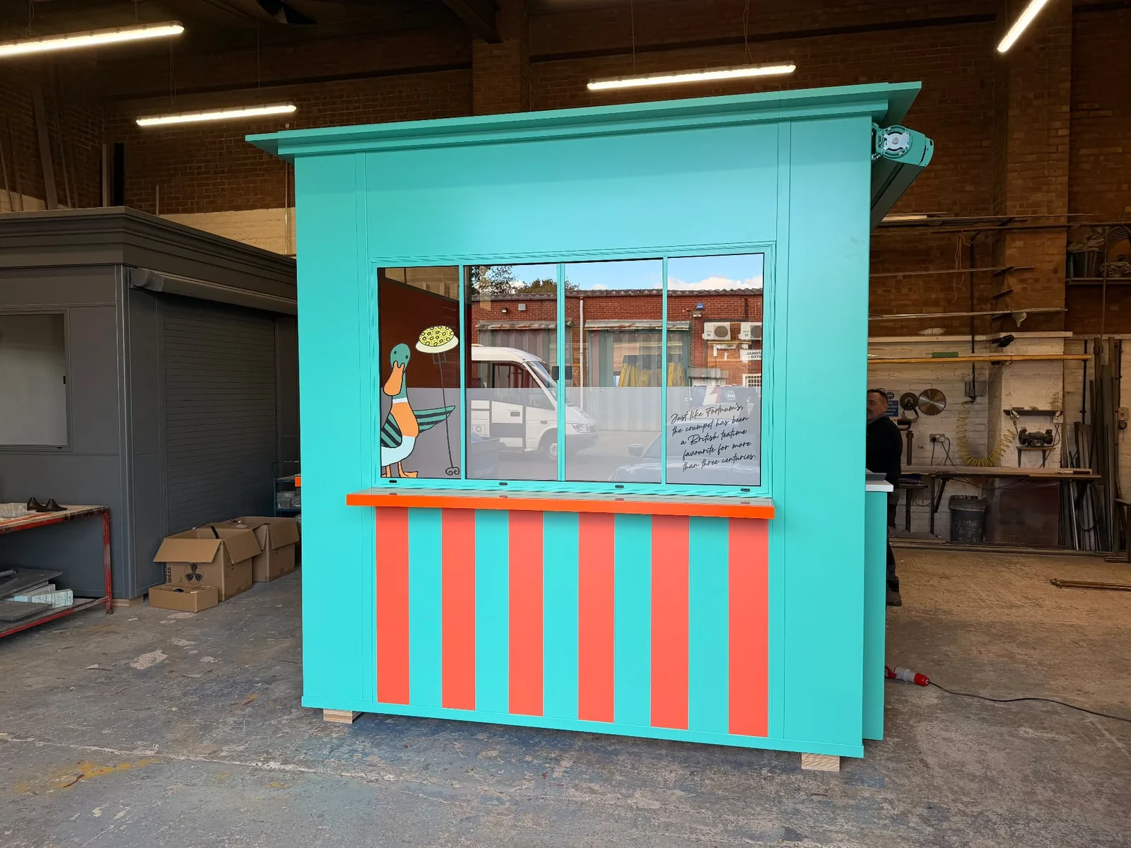 Turquoise kiosk with illustrated window graphics and an orange-and-turquoise striped exterior, at the manufacturer’s premises