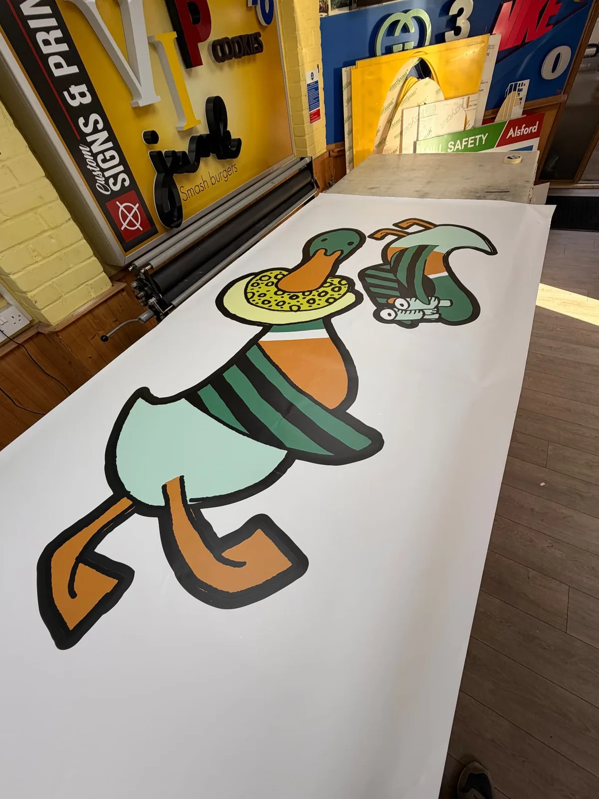 Printed duck illustration graphics laid out on a workbench in the Custom Signs & Print workshop