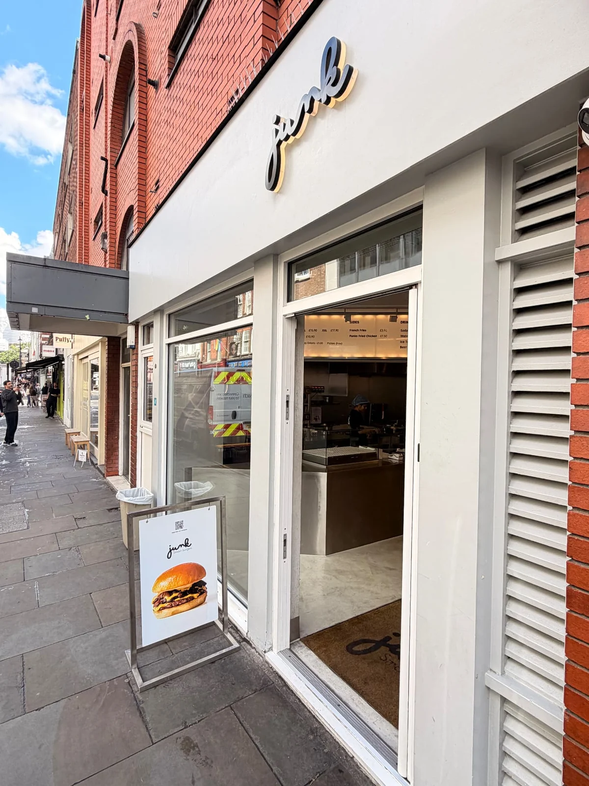 Stainless steel Junk pavement sign and branded entrance mat outside the shopfront, with a Puffy projecting sign further down the street